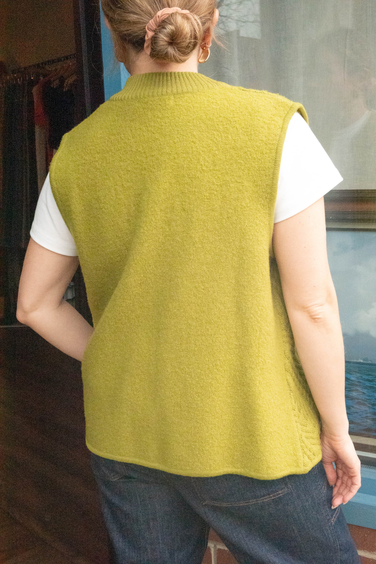 Woman stands with her back turned in front of Petals and Jackets Vintage Shop in West Town Chicago Illinois wearing a vintage green vest from Coldwater Creek, denim oversized pants and a white shirt for her online vintage drops.