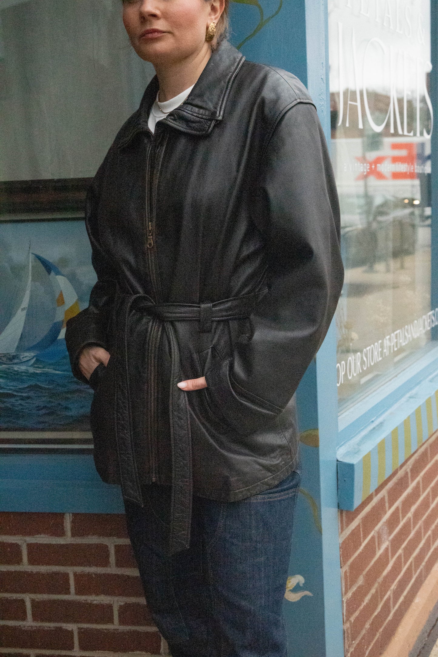 Woman poses in a tied leather jacket with a zipped high neck in front of Vintage Boutique Petals & Jackets in Chicago, Illinois.