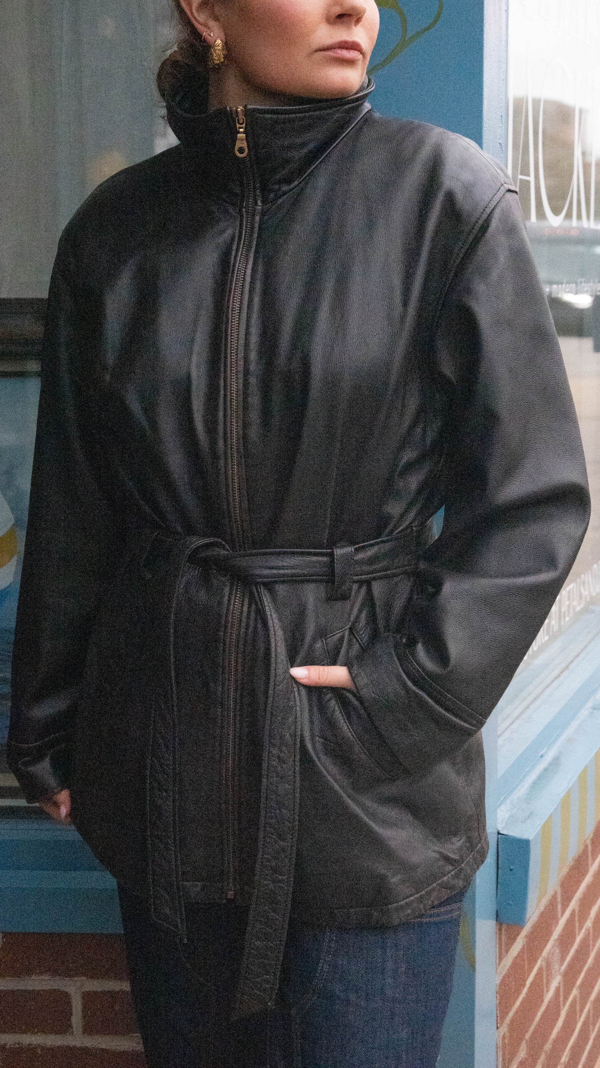 Woman poses in a tied leather jacket with a zipped high neck in front of Vintage Boutique in Chicago, Illinois.